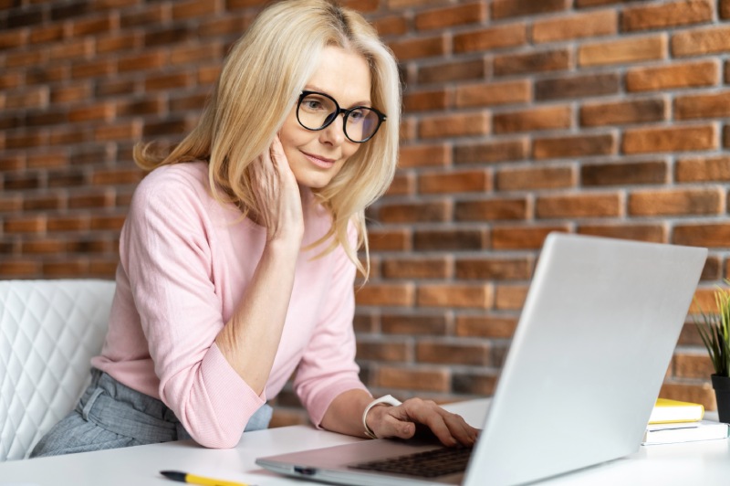 A woman wearing a pink sweater in front of a brick wall while working on a laptop computer