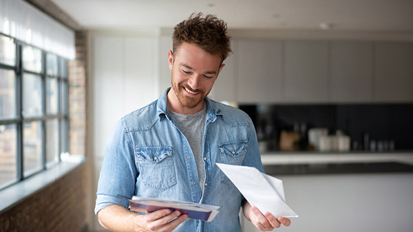 Man in a blue shirt in his kitchen reading a direct mail postcard with interest