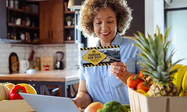 a woman with fresh fruit and vegetables in the kitchen working on her laptop and reading a direct mail postcard