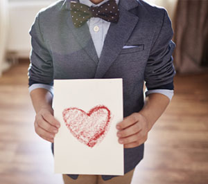 a boy with a bow tie holding up a piece of paper with a heart drawn in crayon