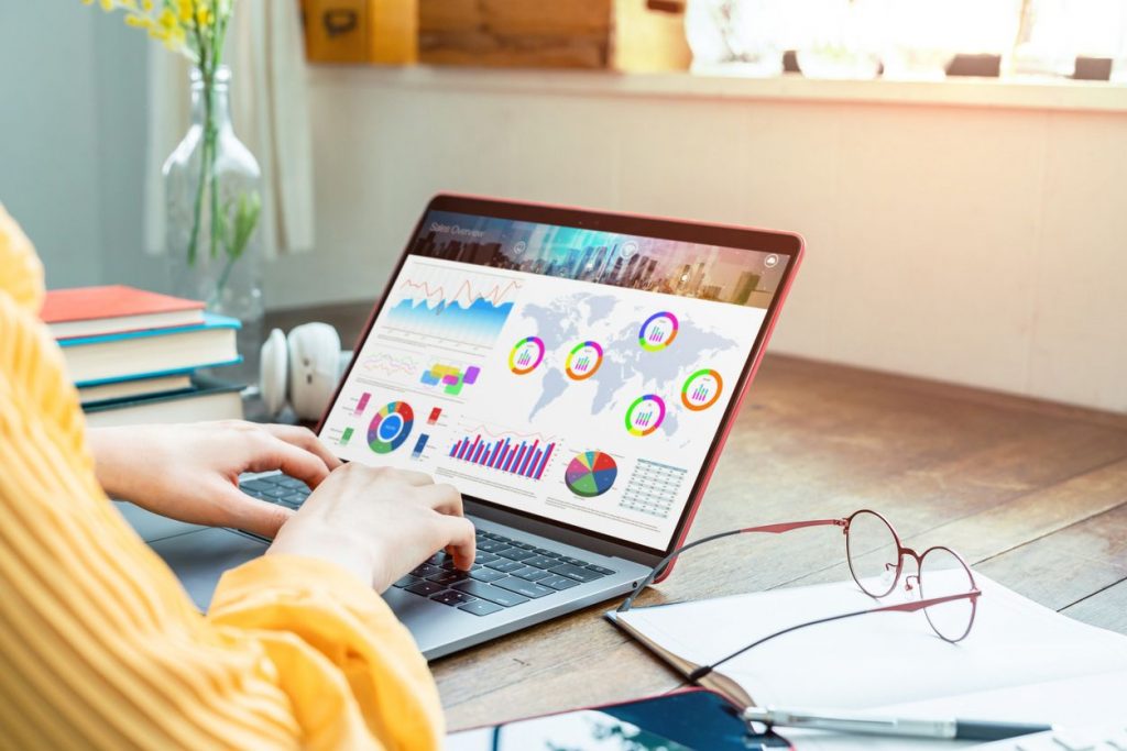 A woman with a yellow long sleeved shirt on working on a laptop with graphs on a computer table with eyeglasses